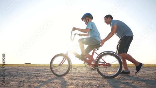 family play in the park. father teaching son to ride a bike. happy family kid dream concept. son learn to ride a bike silhouette. father supporting child son riding bike summer sunset in the park