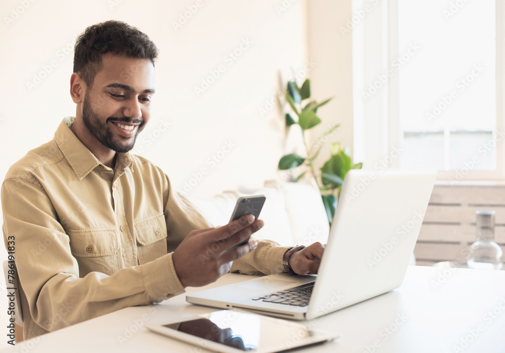 Young handsome man using smart phone and laptop at home. Businessman or student working on computer and texting smartphone. Freelance, education and technology concept Young handsome man using smart phone and laptop at home. Businessman or student working on computer and texting smartphone. Freelance, education and technology concept