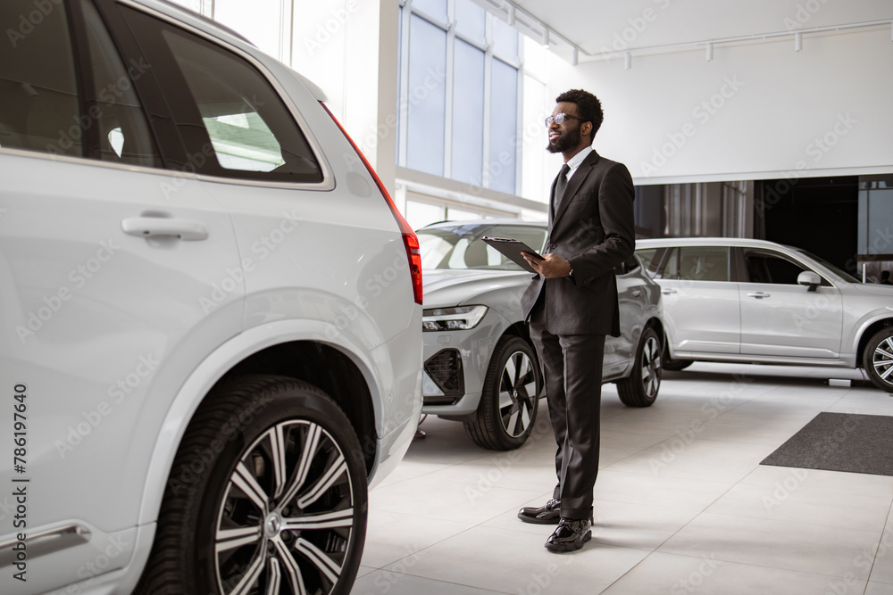 Professional African male vehicle dealer in black suit with clipboard at showroom. Young bearded salesman standing near modern automobile in bright car dealership with panoramic windows.