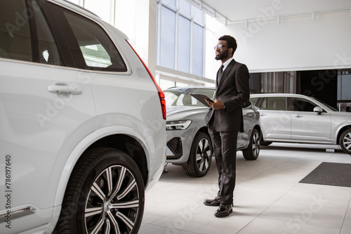 Professional African male vehicle dealer in black suit with clipboard at showroom. Young bearded salesman standing near modern automobile in bright car dealership with panoramic windows. © sofiko14