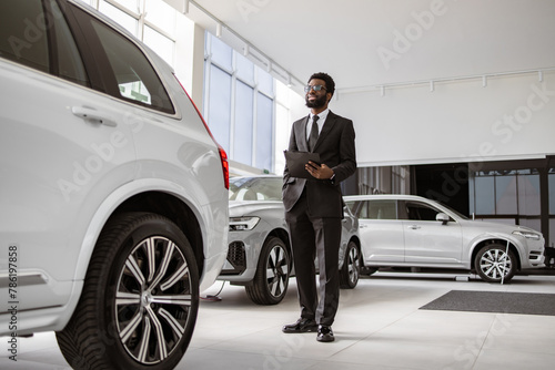 Professional African male vehicle dealer in black suit with clipboard at showroom. Young bearded salesman standing near modern automobile in bright car dealership with panoramic windows. © sofiko14