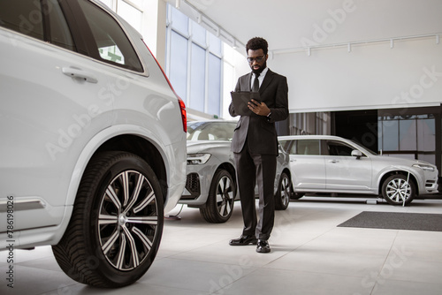 Young bearded salesman standing near modern automobile in bright car dealership with panoramic windows. Professional African male vehicle dealer in black suit with clipboard at showroom. © sofiko14