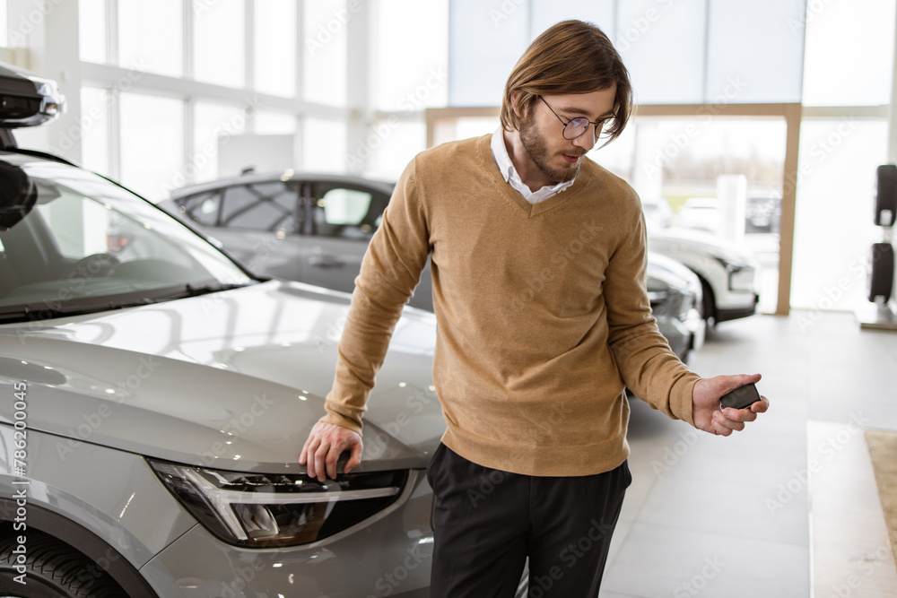 Happy smiling businessman standing near new bought car and holding keys in car dealership. Successful buyer in casual outfit rejoices in a new car. Trading concept.