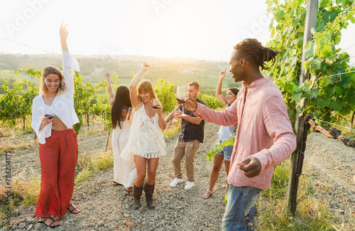 Multiracial friends dancing at summer party inside vineyards - Happy people drinking red wine at coutryside resort - Travel, celebration and tasting event concept - Focus on center blond girl face photo