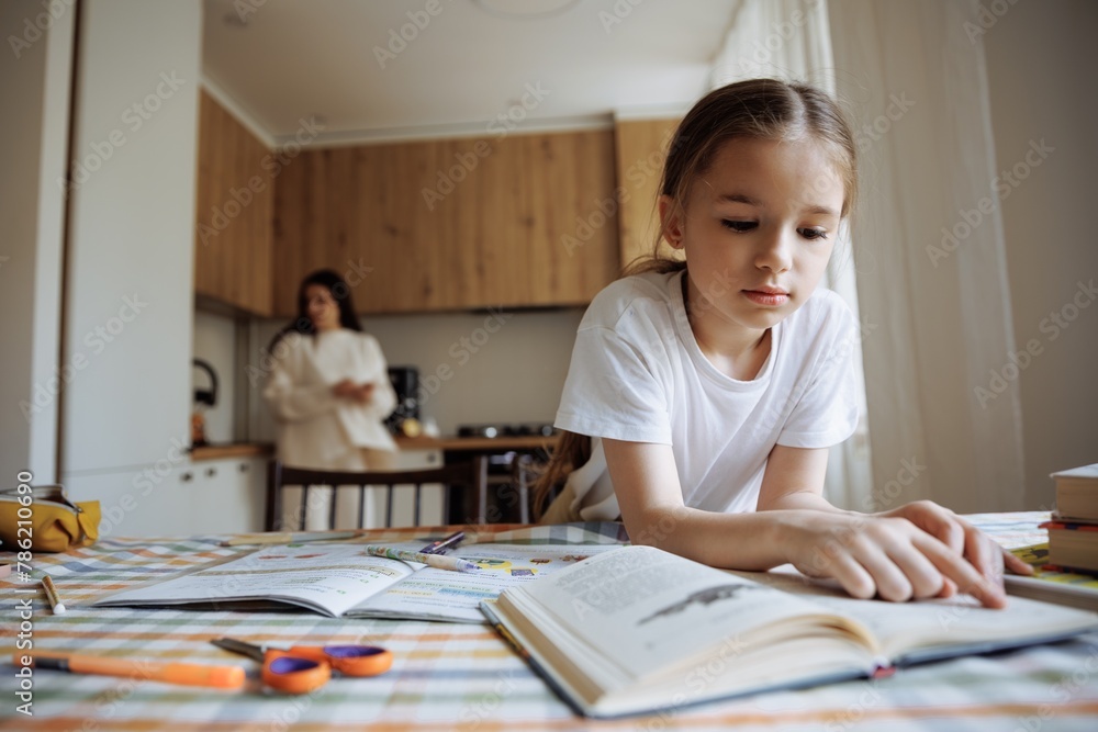 A 6-year-old daughter reads a book and does her homework against the background of her mother cooking in the kitchen A 6-year-old daughter reads a book and does her homework against the background of her mother cooking in the kitchen