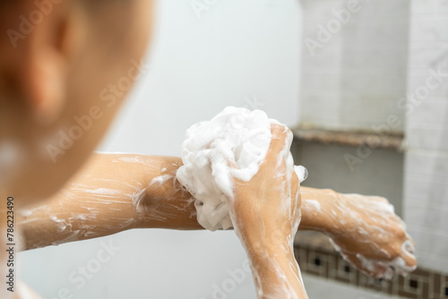 Female shoulder with gel or shampoo foam.Young woman taking shower with moisturizing soap in bathroom. Attractive girl rubbing body skin with white bath puff. Washing arm with white sponge.