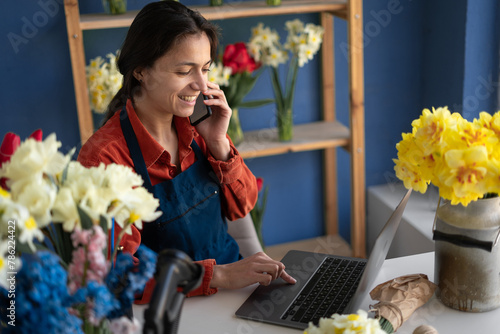 Florist small business flower shop owner. Woman using mobile phone and laptop to take orders for her floral store. Hispanic gardener taking client order during conversation