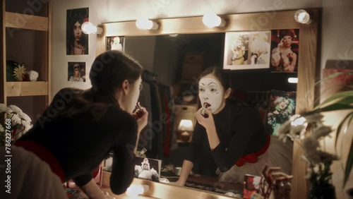 Female mime artist with white face applying red lipstick and looking in mirror white preparing for theatre performance in dressing room photo