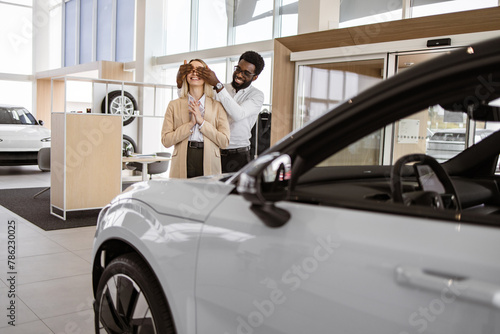 African American boyfriend covering eyes of his Caucasian young girlfriend for surprise in car showroom. Happy husband buying to his loving wife new vehicle at car dealership. photo