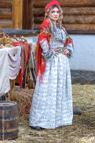 A beautiful Russian girl in traditional Russian clothes and a kokoshnik stands at a table with various national dishes near a wooden house. Festive farewell to winter.