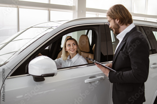 Satisfied happy Caucasian female client customer sitting at steering wheel of new car doing test-drive before buying auto while male shop assistant helping her choose it.