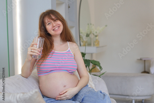 Pregnant Happy woman drink water sit in living room at home on sofa. Young expectant lady have rest at home with glass. Pregnancy, healthcare, healthy lifestyle, looking at camera, stroking belly 