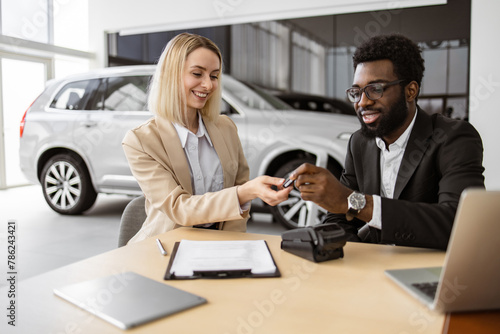 Happy Caucasian young woman sitting at table and receives keys of SUV car from African man salesman in suit in bright showroom. Concept of buying new modern vehicle.