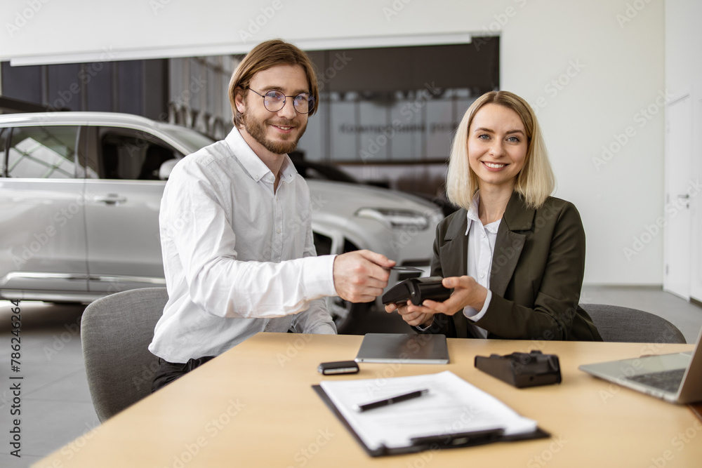 Fototapeta premium Female confident manager helps to male customer make payment for purchase of branded crossover. Happy young man paying with electronic credit card at terminal for purchase of new modern car.
