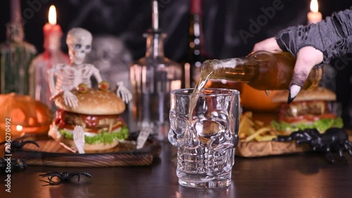 A witch's hand opens a bottle of beer and pours it into a glass in the background, on the table there is a Monster Burger and fries. Halloween party idea. Real time photo