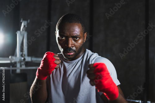 Boxer in gym. Aggressive African man fighter resting after training boxing ready for fight looking at camera. Strong sweated man training punches looking concentrated straight preparing for sparring