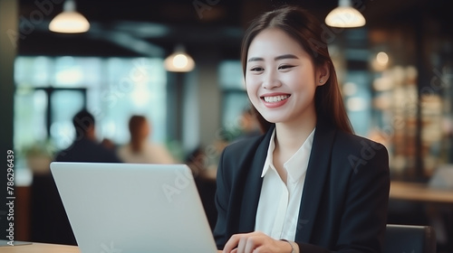 Young Asian woman smiled happily as she utilized technology, working on her laptop in office as a budding entrepreneur in business world. Embodying a professional and successful female entrepreneur. © AK528
