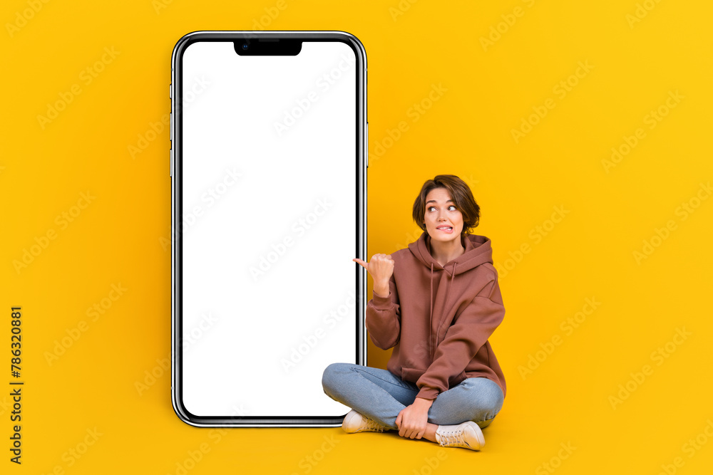 Full length portrait of nice person sit floor biting lips look indicate finger big empty space smart phone screen isolated on yellow color background Full length portrait of nice person sit floor biting lips look indicate finger big empty space smart phone screen isolated on yellow color background