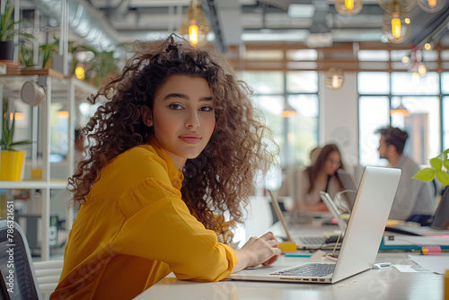 Beautiful Middle Eastern Manager Sitting at a Desk in Creative Office. Young Stylish Female with Curly Hair Using Laptop Computer in Marketing Agency. Colleagues Working in the Background photo