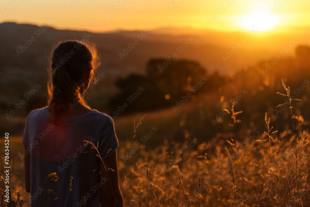 Back view of a woman enjoying a serene sunset in a field, with warm golden light on the horizon