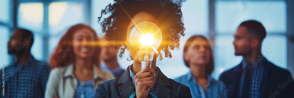 Individual holding a lighted bulb in front, concept of idea creation with peers in background Individual holding a lighted bulb in front, concept of idea creation with peers in background