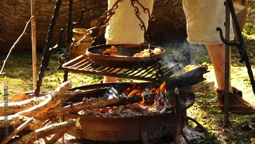 Medieval street kitchen. Man stirns pork meat in cast -iron pans. Suspended metal hearth. photo