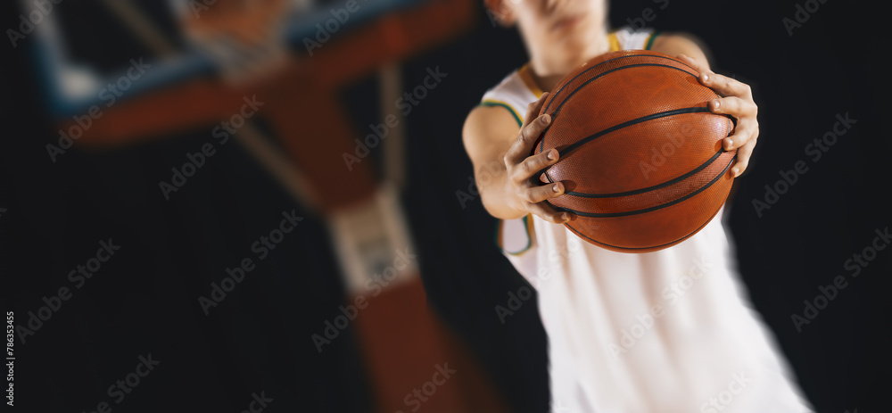 Fototapeta premium Young basketball player with classic ball. Basket in background. Junior level basketball player holding game ball. Basketball training session for kids