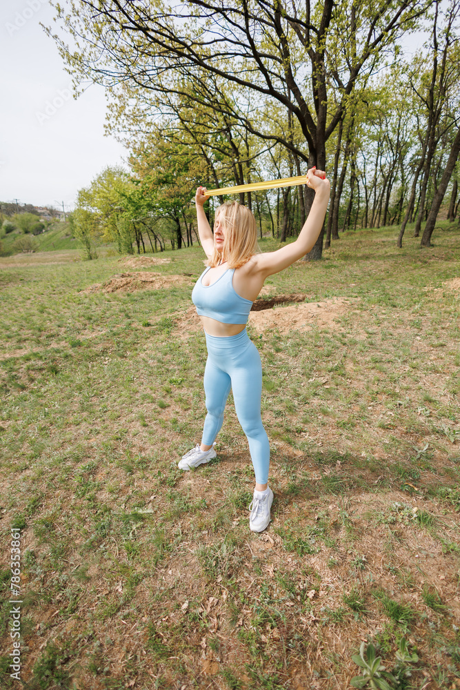 Athletic girl working out in the park stretching a rubber band over her head. Beautiful blonde Caucasian woman in blue tight tracksuit. Blonde girl at an outdoor training session