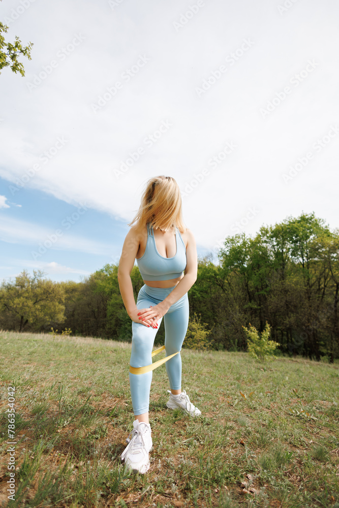 Girl exercising in the park doing inclines with a rubber band on her legs. Beautiful blonde Caucasian woman in blue tight tracksuit. Blonde girl at an outdoor training session Girl exercising in the park doing inclines with a rubber band on her legs. Beautiful blonde Caucasian woman in blue tight tracksuit. Blonde girl at an outdoor training session