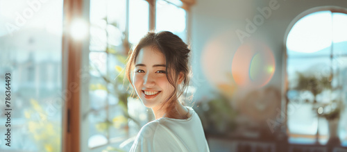 明るく美しいボケの、朝の陽ざしが入る窓辺で優しく笑う20代のアジア人女性