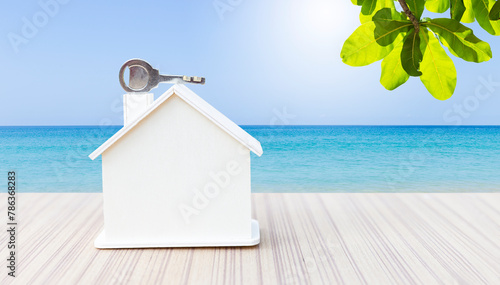 Wooden house model with key over tropical beach background, outdoor day light, real estate business concept, property investment