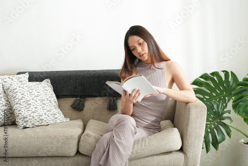 Young beautiful asian woman reading a book at home. Brunette female and rough coated dog sitting on textile couch. White wall background, copy space