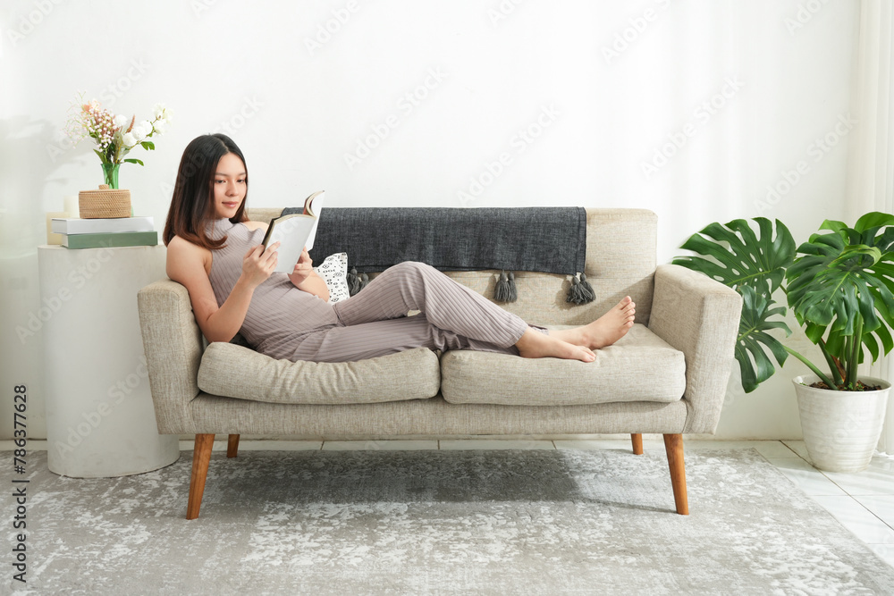 Young beautiful asian woman reading a book at home. Brunette female and rough coated dog sitting on textile couch. White wall background, copy space - obrazy, fototapety, plakaty 