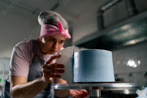 close up female baker in a professional kitchen distributes blue cream onto a sponge cake with a spatula