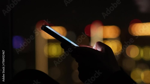 Close-up of female hands holding mobile phone on the background of night city lights. Unrecognizable woman types text messages while sitting on the windowsill in the dark room.