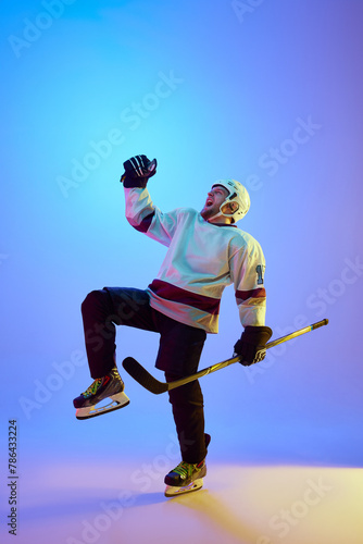 Man, hockey player in helmet, uniform and stick expressing emotions of win against gradient blue background in neon light. Concept of professional sport, competition, game, tournament