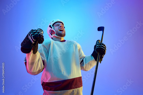 Photography Man, hockey player in helmet, uniform and stick expressing emotions of win against gradient blue background in neon light