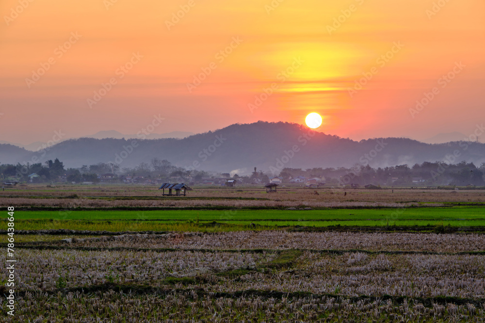 colorful vivid sunset sky from the viewpoint of the roadside of a cropped rice field with orange round sun at the top of the mountain in the background