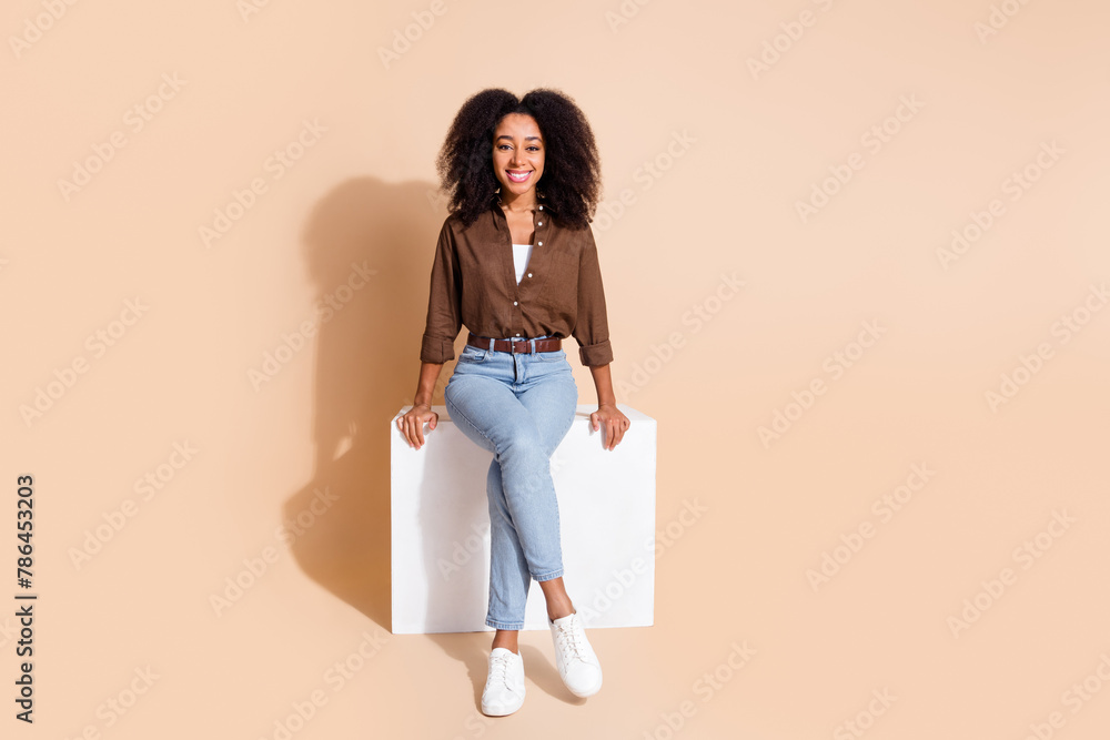 Full size photo of adorable optimistic woman dressed brown shirt denim trosers sit on white cube isolated on beige color background Full size photo of adorable optimistic woman dressed brown shirt denim trosers sit on white cube isolated on beige color background