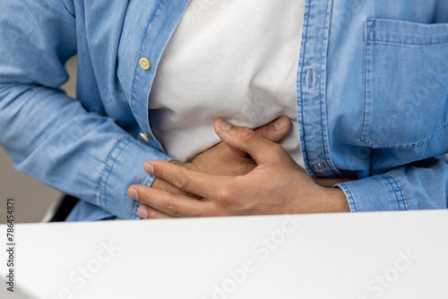 Close-up photo of a body part, hands of a young Latin American man in a blue shirt holding his stomach, feeling and suffering from pain