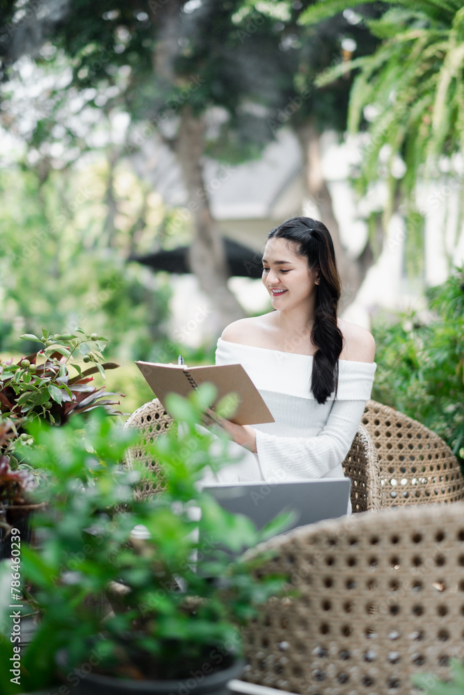 Naklejka premium Smiling freelancer works on a clipboard with a laptop on the table, surrounded by lush garden greenery.