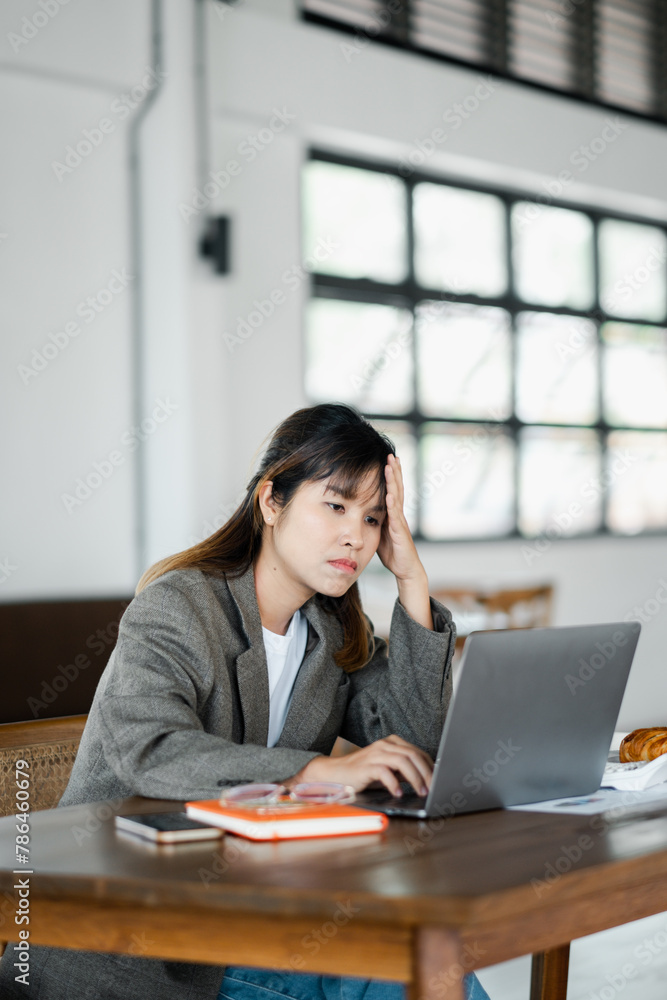 Worried young adult woman working on her laptop in a modern office, showing signs of stress and fatigue.