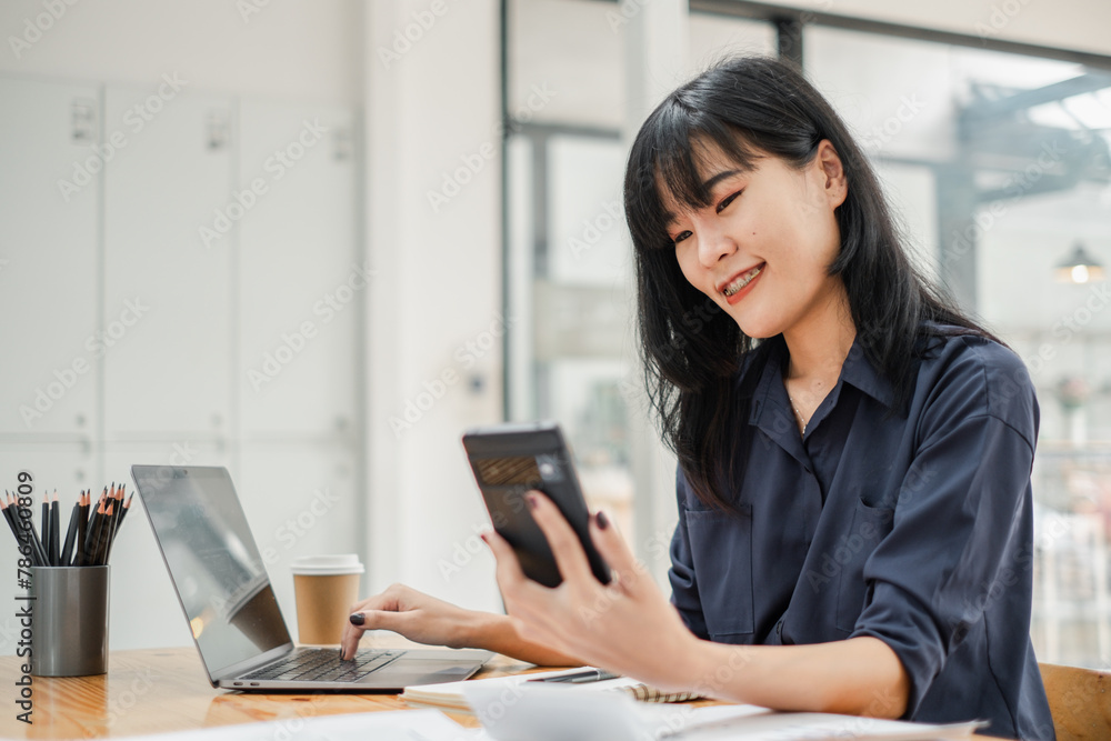 Smiling businesswoman using a smartphone while working on her laptop at a desk with a coffee cup and stationery holder. Smiling businesswoman using a smartphone while working on her laptop at a desk with a coffee cup and stationery holder.
