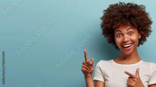 Horizontal shot pleased dark skinned female with Afro haircut, points away with both fore fingers, shows blank space for your promotion, isolated over blue background.. copy space for text. photo