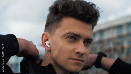 Portrait of a young confident man. He adjusts his jacket and prepares to run. Listens to music in headphones. In the city park