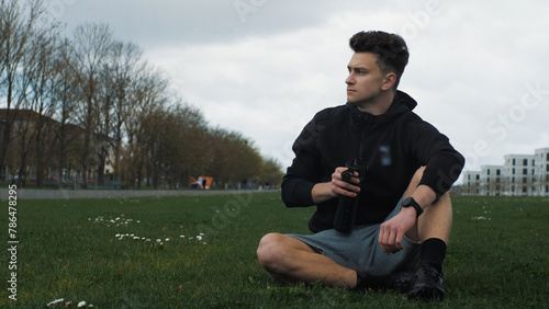 A young man is holding a sports bottle with water. Resting after a run. Sitting in a city park. Healthy Lifestyle