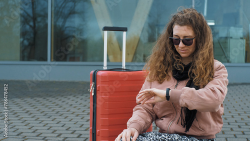 A young woman sits on the asphalt with a red suitcase and waits for transport. Stylish woman in fashionable sunglasses looks at the clock, outdoors. Urban lifestyle