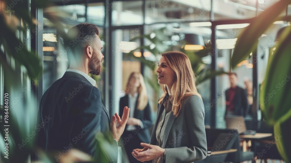 A businesswoman and a male colleague engaged in a conversation, with coworkers in the background, demonstrating teamwork and collaboration in the workplace.  - obrazy, fototapety, plakaty 