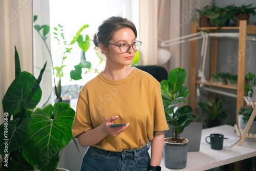 Beautiful young woman working on phone at workstation from home surrounded by indoor plants. Concept of remote work, freelancing, online learning, in the urban jungle.