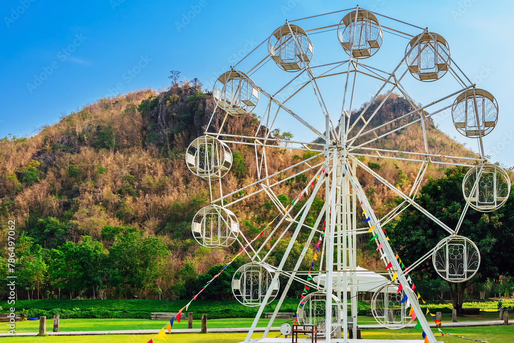 Beautiful scenic with white ferris wheel decorated with lights and small flags for fun and enjoyment of festival or carnival stand on green lawn. Outdoor theme park with beautiful nature as background